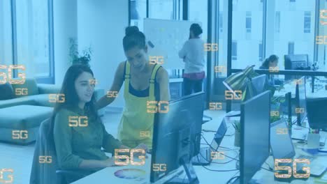 Animation-of-5g-texts-over-diverse-female-coworkers-discussing-reports-on-desktop-in-office
