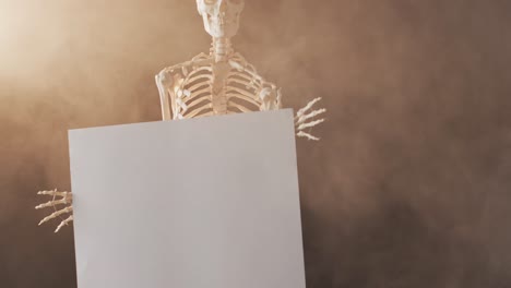 Video-of-close-up-of-halloween-skeleton-holding-white-board-with-copy-space-on-brown-background