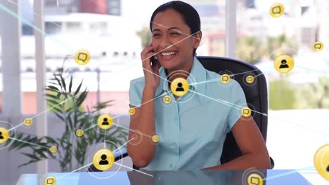 Animation-of-network-of-digital-icons-over-happy-biracial-woman-talking-on-smartphone-at-office