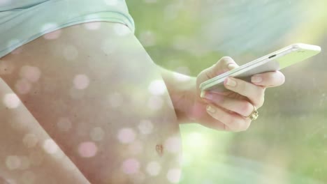 Bokeh-Lichtpunkte-über-Dem-Mittelteil-Einer-Schwangeren-Kaukasischen-Frau,-Die-Ihr-Smartphone-In-Der-Sonne-Benutzt