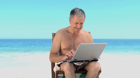 Mature-man-using-a-laptop-sitting-on-beach-chairs