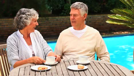 Couple-sitting-chatting-over-coffee-by-a-swimming-pool