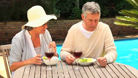 Mature-couple-having-a-meal-with-wine-by-a-swimming-pool