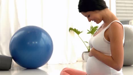 Smiling-pregnant-woman-sitting-on-mat-touching-her-belly
