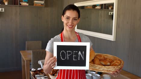 Portrait-of-female-baker-holding-baguettes-and-open-signboard