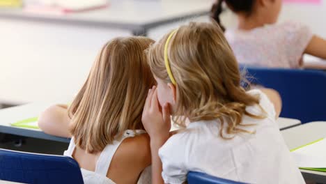 Schoolgirl-whispering-into-her-friend-s-ear-in-classroom