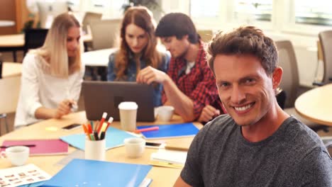 Male-business-executive-smiling-at-camera-while-colleagues-interacting-at-desk