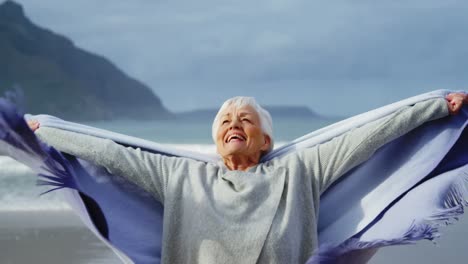 Senior-woman-standing-with-hands-raised-on-beach