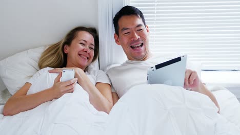 Happy-couple-using-mobile-phone-digital-tablet-on-bed