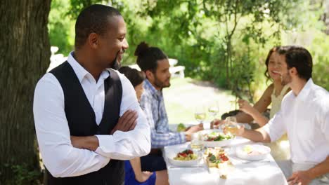 Waiter-standing-with-arms-crossed-while-couples-interacting-in-background