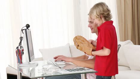 Man-working-with-a-computer-and-son-ready-to-play-baseball