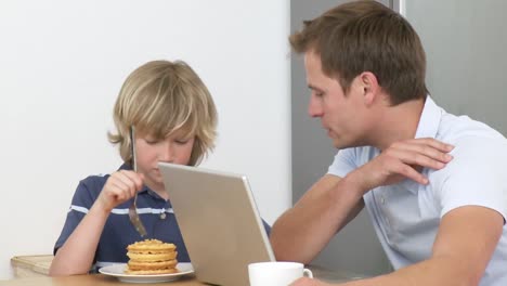 Father-using-a-laptop-and-son-eating-a-sweet-in-the-kitchen