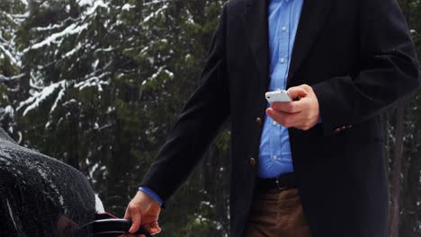 Man-using-mobile-phone-and-charging-the-electric-car-on-a-snowy-day