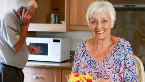 Senior-woman-holding-bowl-of-fruit-while-man-talking-on-mobile-phone-in-kitchen