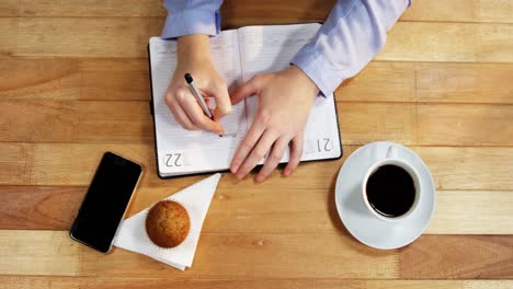 Hand-of-businesswoman-writing-on-diary-with-cupcake,-coffee-and-mobile-phone-on-table