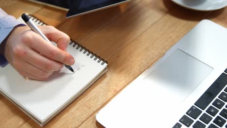 Hand-Einer-Geschäftsfrau,-Die-Am-Schreibtisch-Auf-Ein-Notebook-Schreibt,-Mit-Digitalem-Tablet-Und-Laptop-Auf-Dem-Tisch