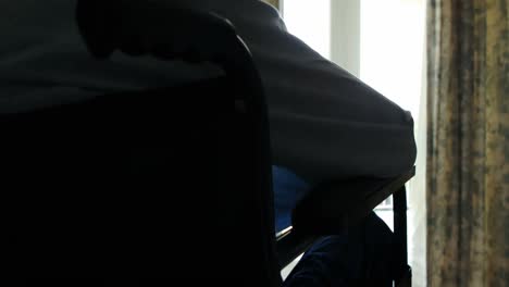 Senior-man-sitting-in-wheelchair-looking-through-window-in-bedroom