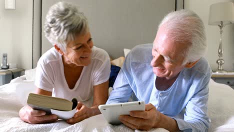 Senior-man-using-digital-tablet-while-woman-reading-a-book-on-bed
