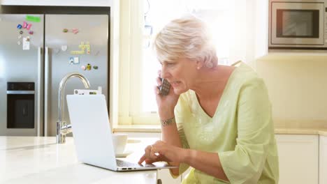 Senior-woman-talking-on-mobile-phone-while-using-laptop-in-kitchen