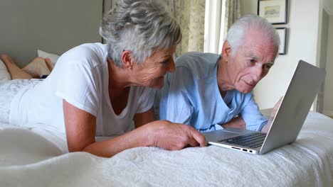Smiling-senior-couple-using-laptop-on-bed-in-bedroom