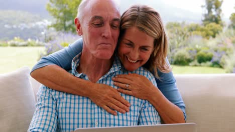 Lächelnde-ältere-Frau-Umarmt-Einen-Mann-Im-Wohnzimmer,-Während-Sie-Einen-Laptop-Benutzt