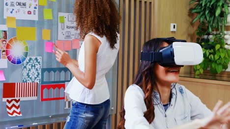 Female-executive-using-virtual-reality-headset-and-colleague-looking-at-sticky-notes