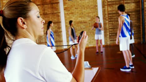 Female-coach-looking-at-stopwatch-while-students-playing-in-basketball-court
