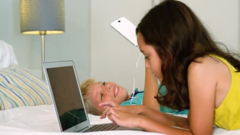 Siblings-sitting-back-to-back-and-using-digital-tablet,-laptop-in-bedroom