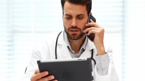 Male-doctor-talking-on-mobile-phone-while-using-digital-tablet
