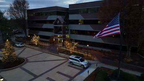 Aerial-orbit-shot-of-waving-American-flag-with-decorated-Christmas-trees-in-front-of-business-park-buildings-in-USA