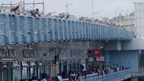 Panoramic-view-of-people-fishing-at-Galata-Bridge-with-famous-Turkish-seafood-restaurant-in-view