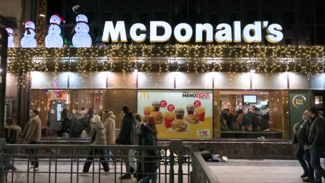 Los-Peatones-Pasan-Por-La-Cadena-De-Restaurantes-De-Hamburguesas-De-Comida-Rápida-Multinacional-Estadounidense,-McDonald&#39;s,-Decorada-Con-Luces-Navideñas