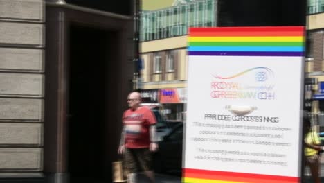 Royal-borough-of-Greenwich,-pan-shot-of-traffic-light-with-sign-about-pride-month,-gay-pride,-with-rainbow-pedestrian-crossing-in-blurred-background,-day,-real-time