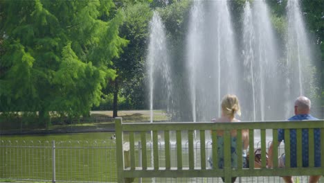 Una-Pareja-De-Ancianos-Sentados-En-Un-Banco-Del-Parque-Frente-A-Una-Fuente-De-Agua-En-El-Verano