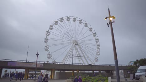 Ein-Riesenrad-Mit-Blinkenden-Lichtern-An-Einem-Bewölkten-Nachmittag,-Im-Vordergrund-Laufen-Menschen