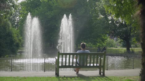 Un-Hombre-Sentado-En-Un-Banco-Del-Parque-Frente-A-Una-Fuente-De-Agua-En-Leamington-Spa-En-El-Verano