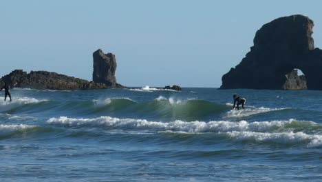 Surfistas-En-Trajes-De-Neopreno-Montando-Una-Ola-A-Lo-Largo-De-Toda-La-Pantalla-Con-Dolor-De-Roca-En-El-Fondo-Panorámica-De-Derecha-A-Izquierda