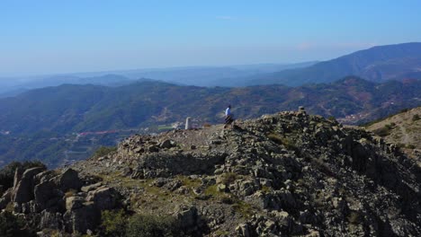 aerial-drone-shot-circling-man-stood-at-the-top-of-a-mountain,-revealing-picturesque-mountain-range-in-the-background