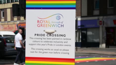 Royal-borough-of-Greenwich,-tilt-shot-of-traffic-light-with-sign-about-pride-month,-gay-pride,-with-rainbow-pedestrian-crossing-in-blurred-background,-day