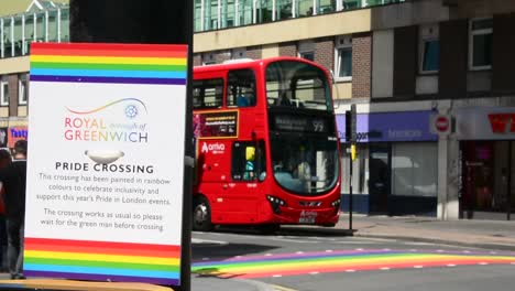 Barrio-Real-De-Greenwich,-Plano-De-Semáforo-A-La-Altura-De-Los-Ojos-Con-Un-Cartel-Sobre-El-Mes-Del-Orgullo,-Orgullo-Gay,-Con-Cruce-Peatonal-Arcoíris-Y-Un-Icónico-Autobús-Rojo-De-Dos-Pisos-Con-Fondo-Borroso,-Día