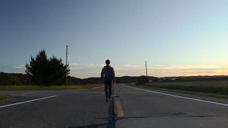 Ein-Mann-Geht-In-Der-Abenddämmerung-Langsam-Mitten-Auf-Einer-Ländlichen,-Rissigen-Asphaltstraße-In-Die-Ferne