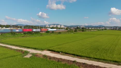 Vista-Aérea-De-Un-Tren-De-Carga-Que-Transporta-Contenedores-Mientras-Cruza-El-Campo-Con-Campos-De-Arroz-En-Primer-Plano-Y-Edificios-De-Apartamentos-Al-Fondo.