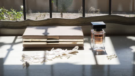 Sun-shining-trough-Glass-Perfume-Bottle-on-white-table-next-to-a-purse,-flowers-and-earrings