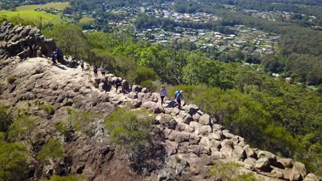 Toma-Aérea-De-Seguimiento-Por-Drones-De-Un-Grupo-De-Personas-Caminando-Por-Una-Montaña-Rocosa,-En-Un-Día-Soleado,-En-El-Parque-Nacional-Glass-House-Mountains,-En-Queensland,-Australia