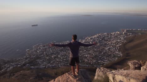 Hombre-Levantando-Las-Manos-Sobre-La-Espectacular-Vista-De-Ciudad-Del-Cabo-En-La-Cima-De-La-Montaña-Cabeza-De-León-En-Cámara-Lenta