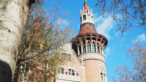 Mirando-Hacia-Arriba,-árbol-Que-Revela-Una-Vista-Panorámica-Del-Diseño-Exterior-De-La-Torre-Del-Parque-Guell-En-Barcelona,-Cielo-Azul-Nublado-En-El-Fondo