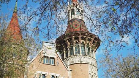 Establishing-UpShot,-Tree-branches-and-green-leaves-waving,-Revealing-Park-Guell-Entrance-tower-in-Barcelona,-Blue-Sky-In-the-background