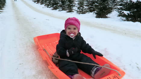 Una-Niña-Pequeña-Con-Un-Sombrero-Rosa-Montando-Un-Trineo-Naranja-Por-Una-Pequeña-Colina-Nevada-En-Una-Granja-De-árboles-De-Navidad