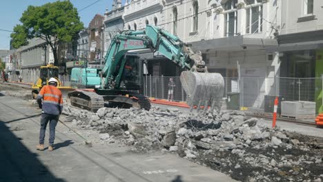 Trabajador-De-Plano-Amplio-Barriendo-Mientras-La-Excavadora-Rompe-El-Concreto-De-La-Carretera-Para-Reemplazar-Las-Líneas-Del-Tranvía,-Con-Una-Topadora-Detrás