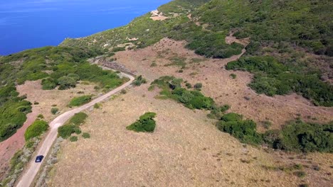 Toma-Aérea-De-Drones,-Bajando-Y-Siguiendo-Un-Auto-Negro-En-La-Carretera-Costera-En-Un-Paisaje-Mediterráneo-En-Cerdeña,-Italia.